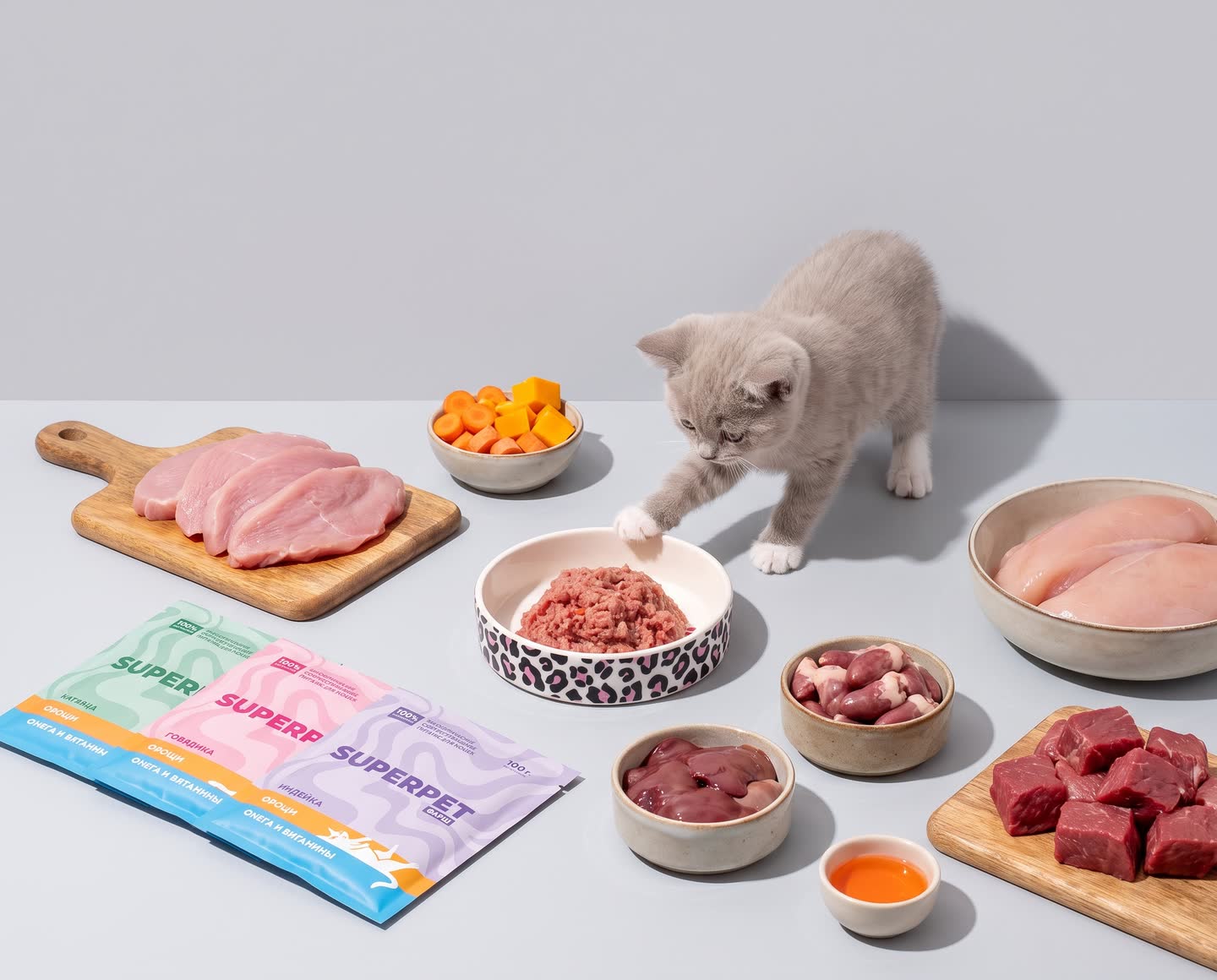 Котёнок тянется лапой к миске с сырым мясом рядом с упаковками SUPERPET